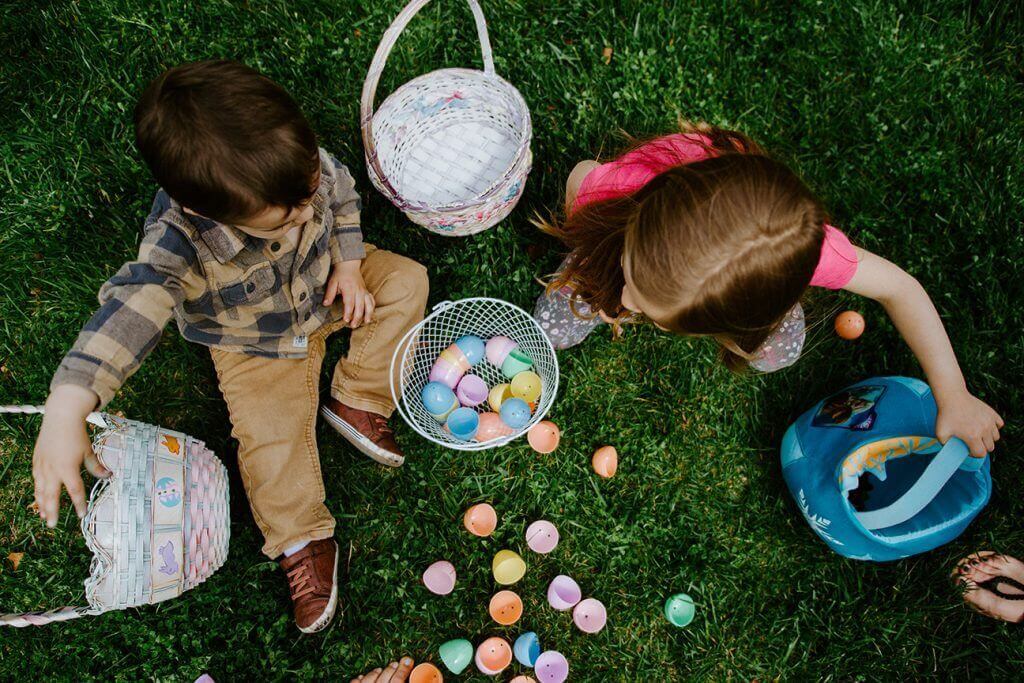 Two young children sitting on grass with Easter egg baskets, surrounded by chocolate Easter eggs